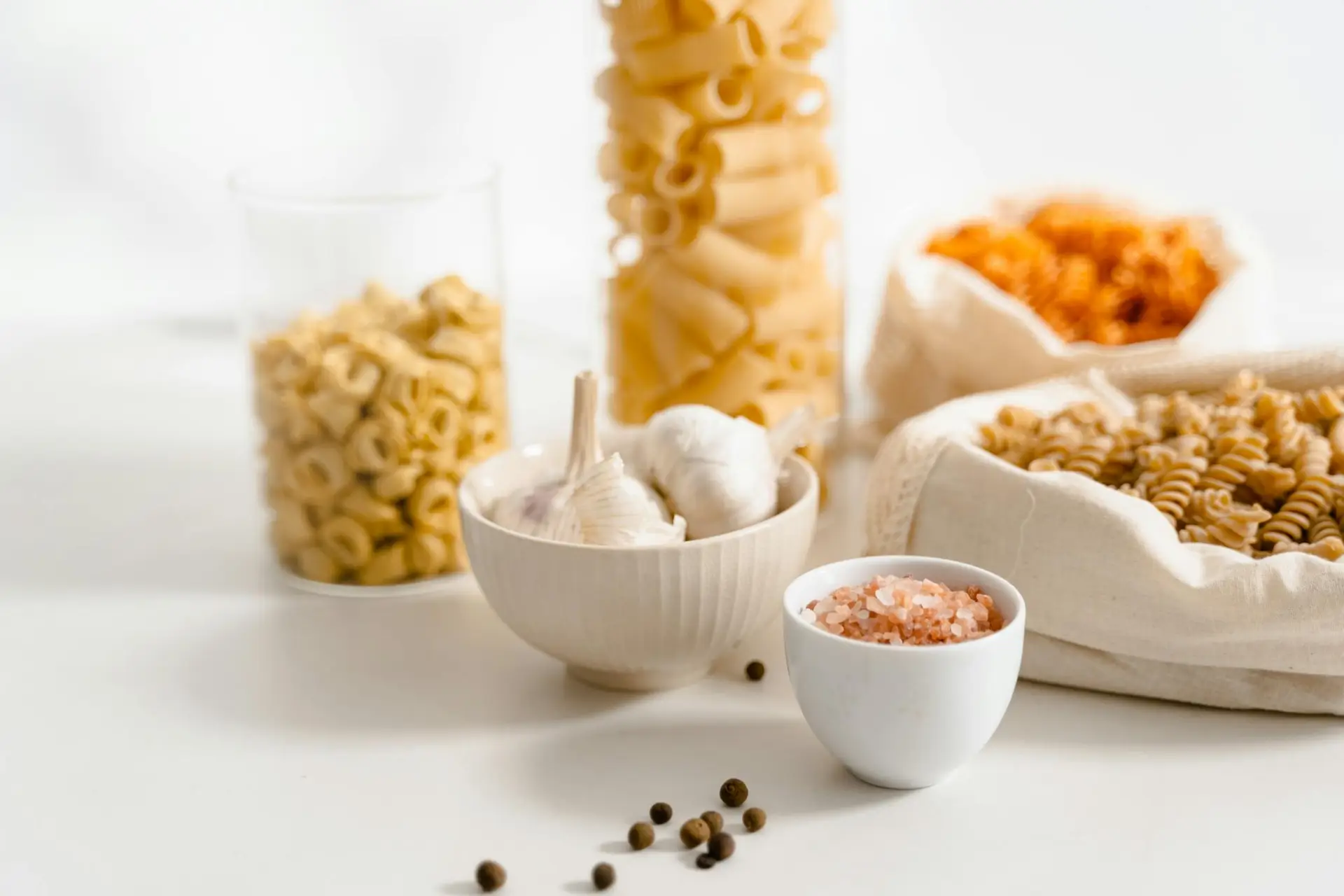Assorted raw pasta with garlic, pink salt, and pepper on a white surface.